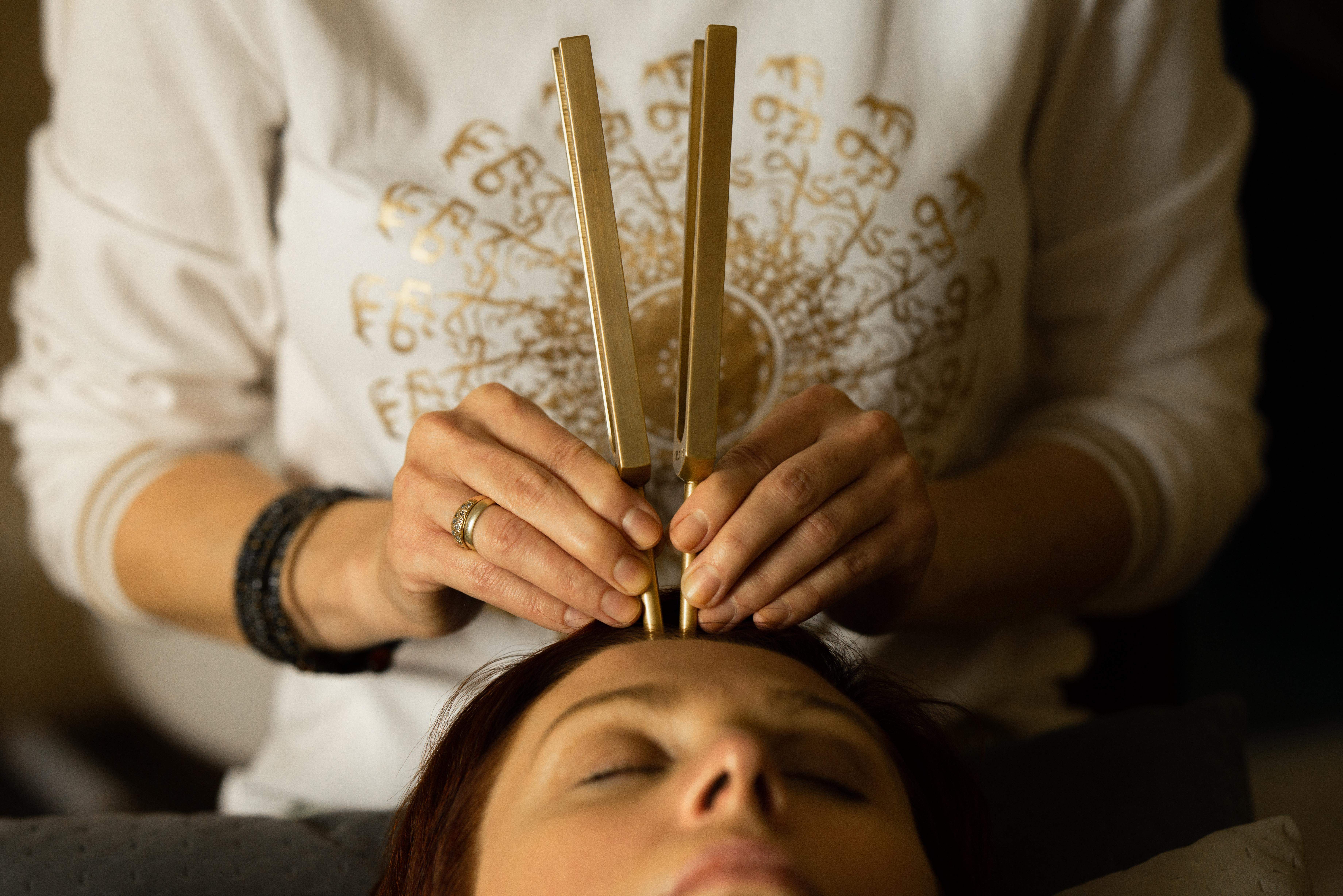 Kamertony trzymana w rękach pani. Dwa kamertony mosiężne postawione na punkcie  trzeciego oka na twarzy kobiety która leży na plecach.